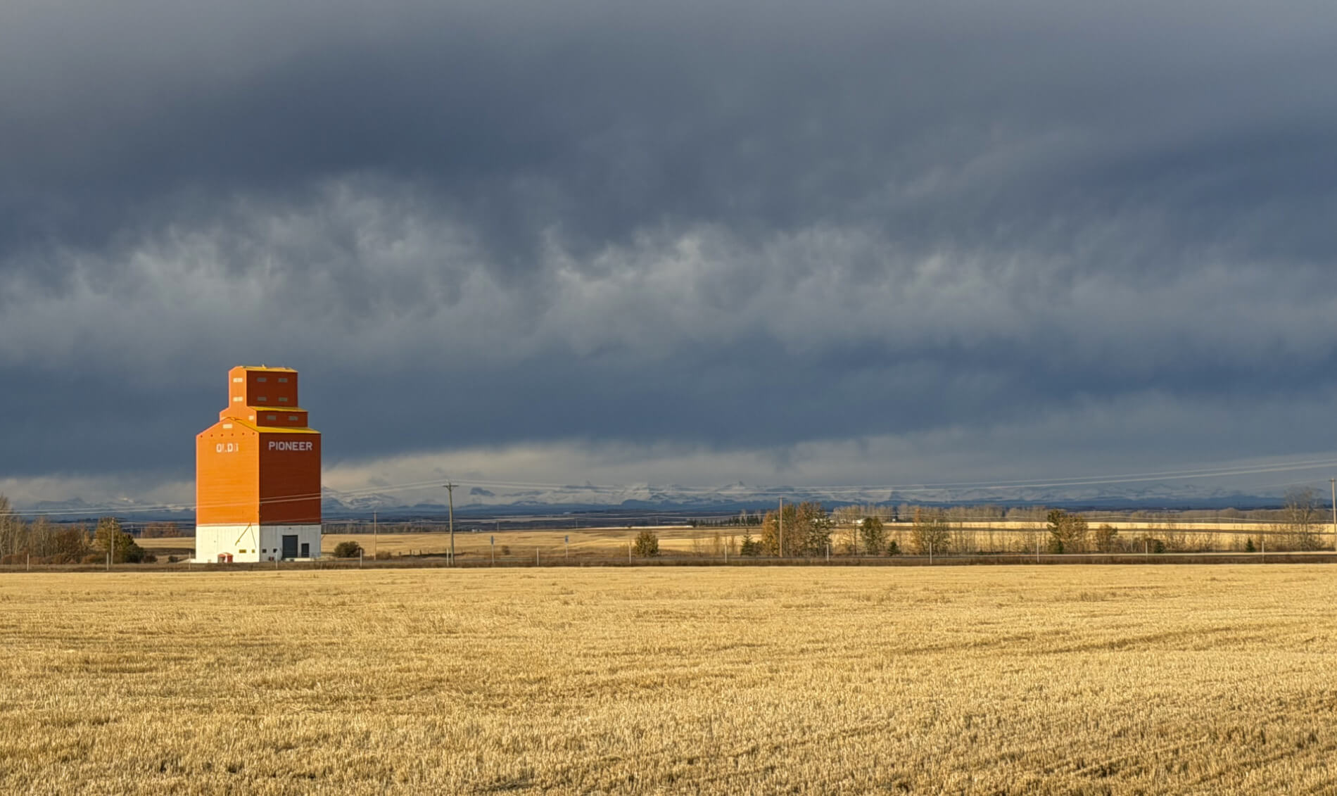 A big red grain bin with Olds written on it, sitting in a prairie field with a beautiful mountain range behind it